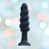 The VeDO Twist Plug Vibrating Butt Plug - Black, a spiral-shaped silicone anal plug with a flared base and 10 vibration modes, is centered against a blurred background featuring soft, out-of-focus circular lights in various colors.